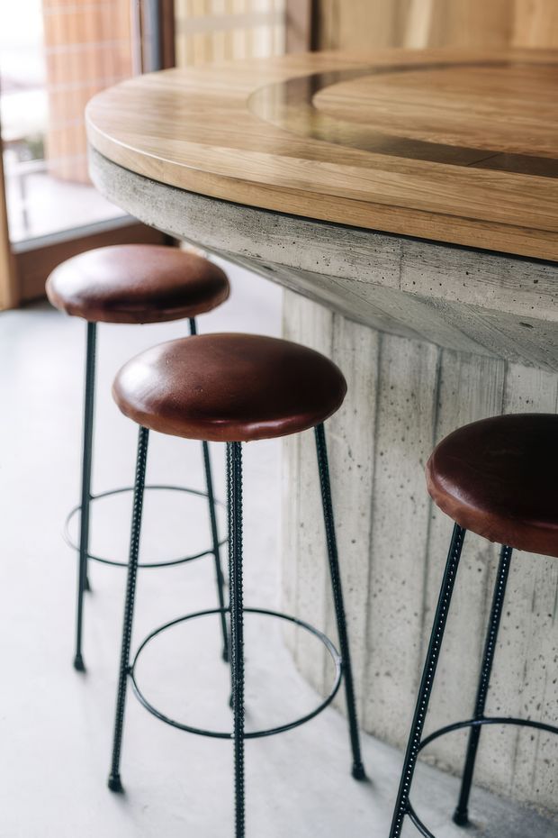 Close-up interior detail of a curved bar or kitchen island with a thick natural timber benchtop set over a raw concrete base. Three dark brown leather-topped bar stools with slim black metal rebar-style legs sit on a pale floor, with glazing and warm timber elements in the background. The image highlights material contrast between timber, concrete, leather, and metal.