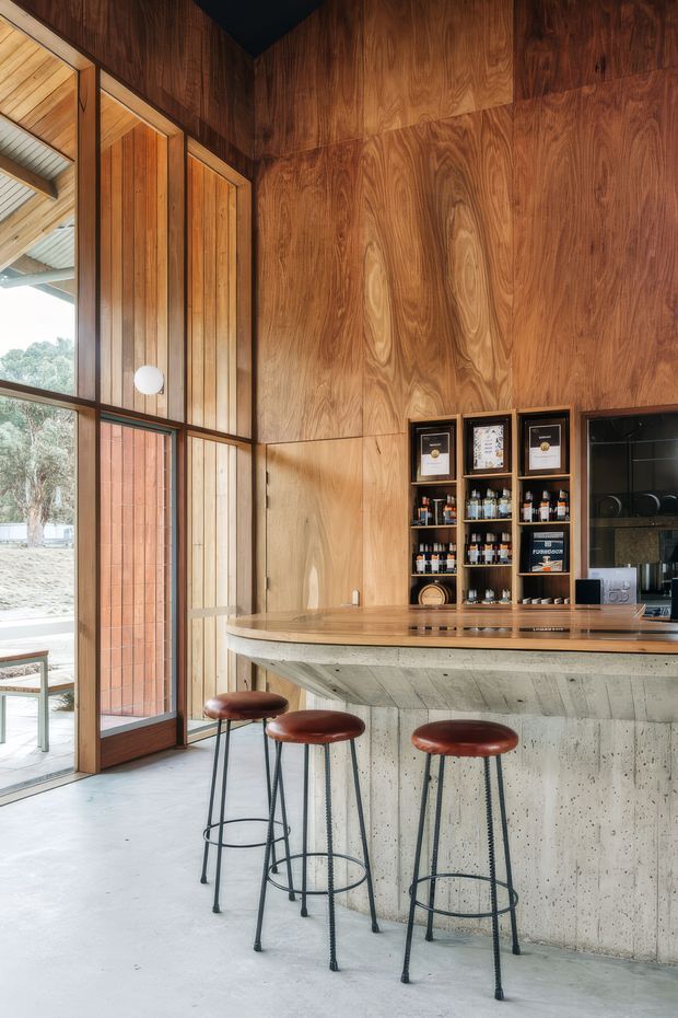 A warm, timber-lined bar or café interior with a polished timber counter, integrated shelving stocked with bottles and framed certificates, and a textured concrete bar front. Three slim metal bar stools with brown leather seats sit at the counter. Full-height glazed timber doors and windows bring in daylight and provide views to trees outside. The space has a refined contemporary feel with strong natural-material detailing.