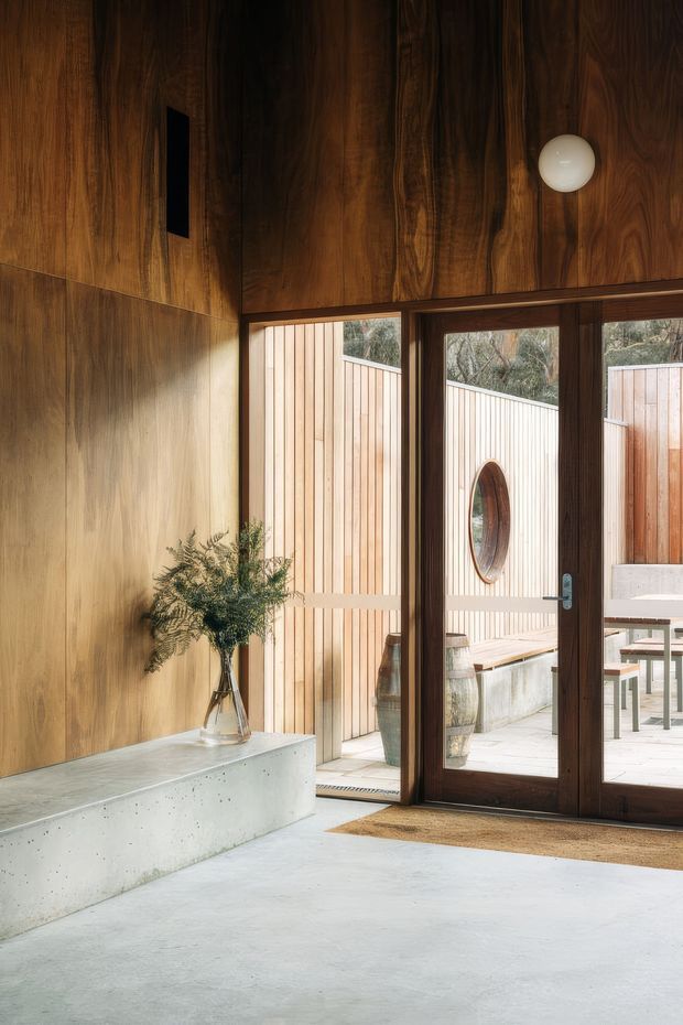 Warm contemporary interior entry space with extensive dark timber wall and ceiling panelling, a polished concrete floor, and a built-in concrete bench. Full-height timber-framed glazed doors open to a private timber-clad courtyard, while a spherical wall light and a small vase arrangement add restrained detail.
