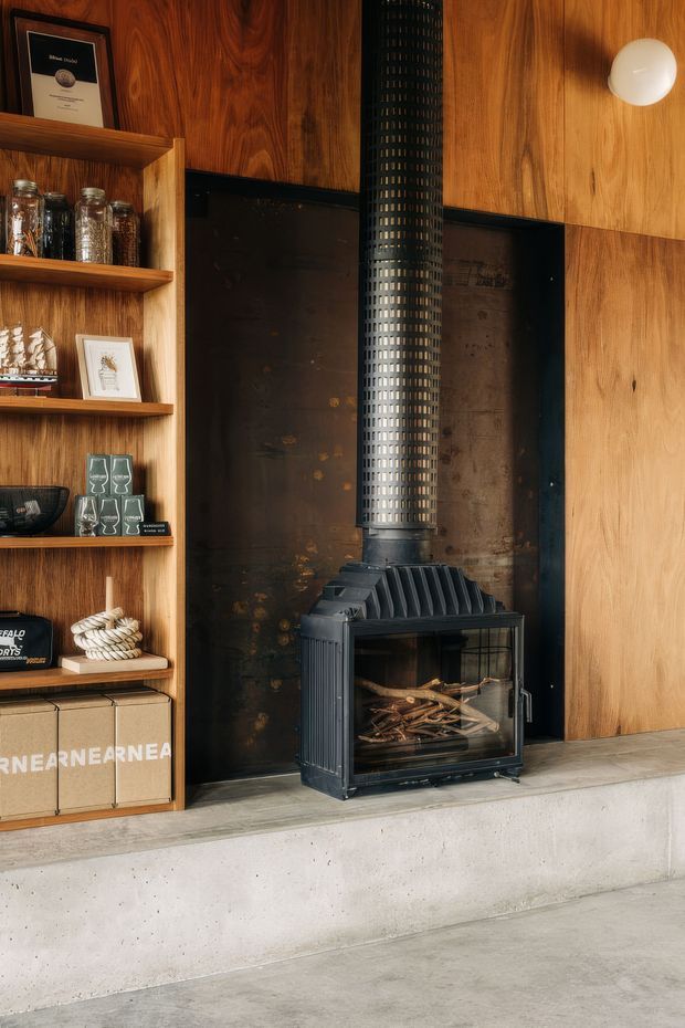 A close-up interior view of a built-in fireplace installation. A black cast-iron wood-burning stove with a tall flue stands against a dark metal hearth surround, framed by warm timber wall panelling and open timber shelving displaying glassware and small decor. The fireplace is set above a light grey concrete plinth and floor, with a simple round wall light on the right. The composition has a warm, refined industrial character with strong timber and black metal detailing.