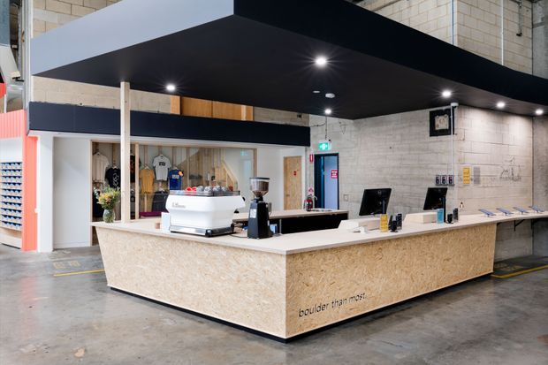 A modern commercial café or reception counter in an industrial warehouse-like interior. The large island counter has a light wood-fibre finish, white solid-surface countertop, espresso machine and register stations. A black suspended canopy with recessed lighting defines the service area, while pale concrete block walls, exposed industrial finishes and polished concrete flooring surround it. Merchandise and a retail display are visible behind the counter.