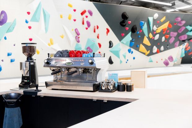 Bright contemporary commercial café interior with a professional espresso machine and coffee grinder on a long light-coloured counter. Black built-in cabinetry forms the base, while the rear wall features a bold multicoloured rock-climbing wall with geometric climbing holds. Exposed ceiling services and linear lighting reinforce the casual, high-energy hospitality setting.