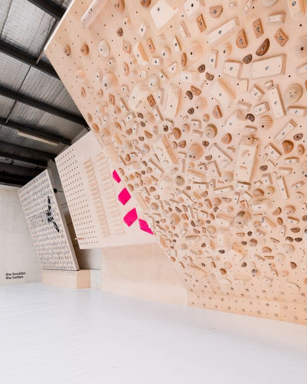 Interior view of a large indoor climbing gym featuring an extensive pale timber climbing wall with dense arrays of climbing holds and volumes. The wall forms steep and overhanging planes beneath an exposed dark metal and corrugated industrial roof structure. Additional climbing walls and bright pink geometric holds are visible in the background, with a spacious light-coloured floor.