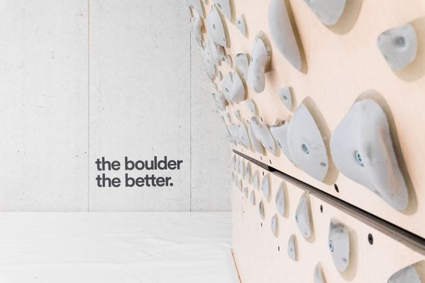 A clean, minimalist indoor climbing wall installation with pale timber panels and numerous light-coloured climbing holds. A smooth light-grey concrete wall and white floor create a bright, restrained setting, with the slogan “the boulder the better.” printed on the wall.