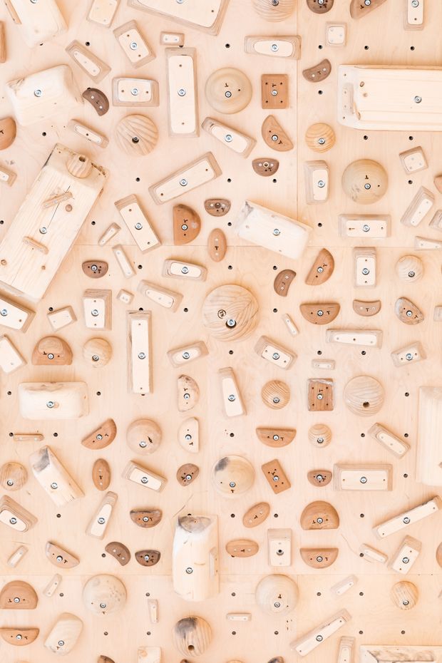 A close-up, top-down view of a pale timber climbing wall densely covered with varied wooden climbing holds in cream, tan, and brown tones. The composition is repetitive and graphic, with many visible bolts and mounting holes; no room context is shown.