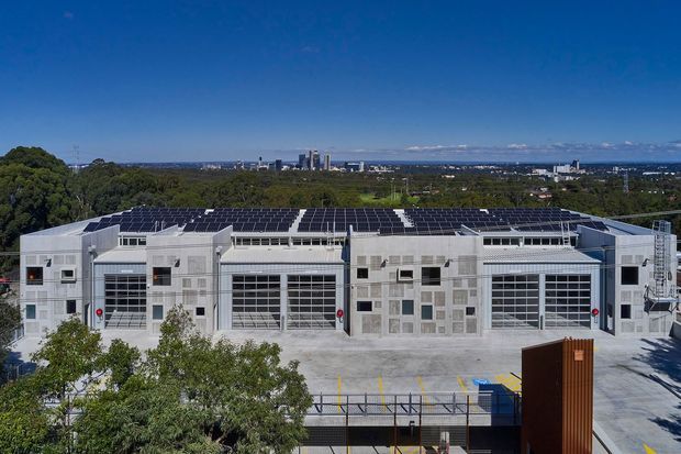Aerial exterior view of a large contemporary light-grey industrial or commercial building with multiple glazed sectional doors, rooftop solar panels, rooftop equipment, and a broad paved forecourt. Dense trees and a distant urban skyline are visible beyond the building.