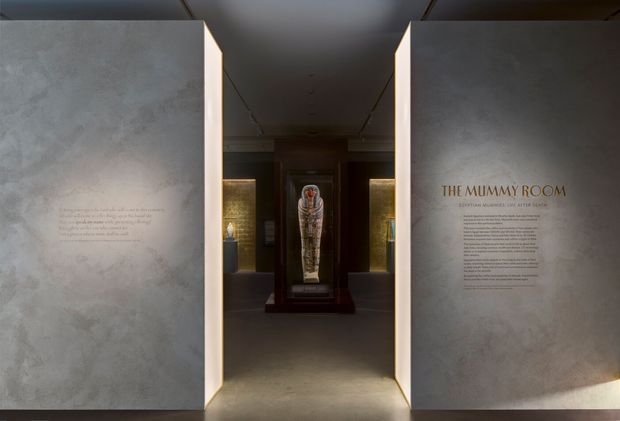 A dimly lit museum or exhibition interior with a central illuminated Egyptian-style mummy display in a dark wood or bronze-toned case. Large pale textured partition walls frame the view, with exhibition text printed on both sides, including the heading “THE MUMMY ROOM.” Recessed and concealed lighting creates a dramatic, focused atmosphere.