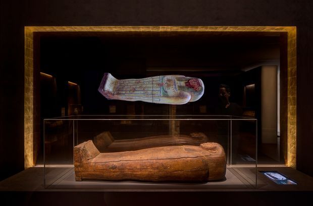 A dark, museum-like exhibition space displaying an ancient wooden sarcophagus or funerary figure inside a clear glass vitrined case, with a suspended illustrated artifact above it. The display is framed by warm, dark timber or bronze-toned architectural trim and dramatic low-level lighting.