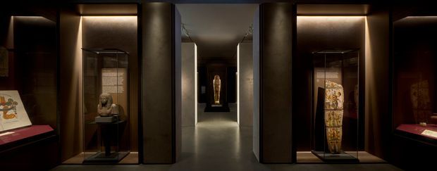 A dark, refined museum or gallery interior with symmetrical illuminated display cases containing sculptures and artefacts. A central corridor leads toward a further spotlighted exhibit, while recessed warm lighting highlights the black and bronze-toned cabinetry and walls.