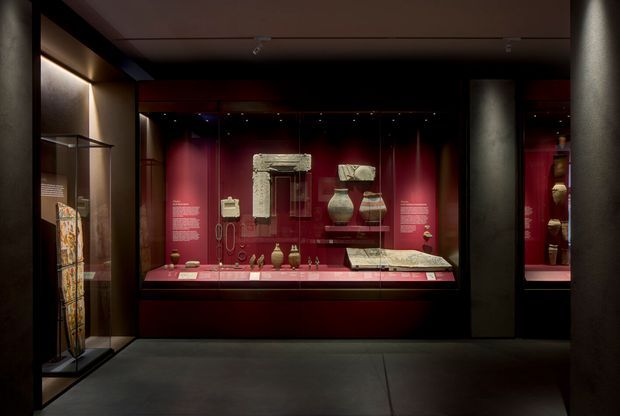 A dimly lit museum or gallery exhibition interior with dark walls, red display niches, glass-fronted vitrines, and illuminated historical artefacts arranged on shelves and plinths. The space has a refined, dramatic presentation with dark flooring, black structural framing, and focused track lighting.