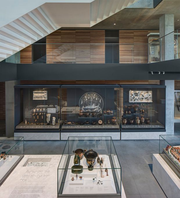Interior view of a contemporary museum or gallery exhibition space with glass display cases containing artifacts and objects. The room features dark charcoal built-in display joinery, concrete structural elements, extensive warm timber wall panelling, polished grey flooring, and a dramatic white stair or folded ceiling structure overhead.