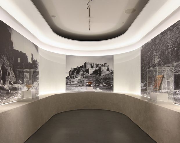 A refined museum or gallery interior with a curved central display plinth, three large monochrome historic landscape murals or photographic panels, and small glass display cases. The space features sculptural curved walls, concealed perimeter lighting, a recessed oval ceiling detail, and a dark neutral floor, creating a quiet, immersive exhibition environment.