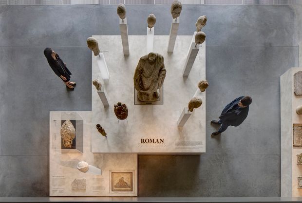 Overhead view of a museum or gallery exhibition featuring a central Roman sculpture on a pale stone display platform, surrounded by elevated sculptural fragments and information panels. Visitors stand on a large dark grey floor surface around the exhibit. The composition is minimal, curated, and institutional, with a muted neutral palette and strong geometric arrangement.