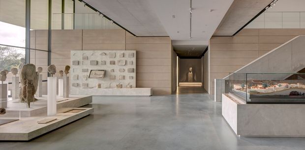 A spacious, minimalist exhibition or gallery interior with polished concrete flooring, pale timber wall panelling, full-height glazing, and a clean white ceiling. Sculptural objects and artefacts are displayed on low plinths and within glass cases, while a broad stair or ramp and a dark central corridor lead deeper into the space. The restrained palette and precise detailing create a refined, museum-like atmosphere.