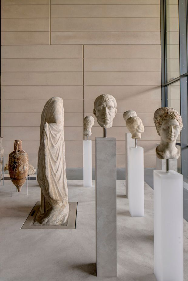A quiet museum or gallery interior displaying classical stone sculptures, busts, and an ancient vessel on tall minimalist plinths. The space features pale beige horizontal wall panelling, a smooth light grey floor, and a large glazed window at right, creating a refined, calm exhibition setting.