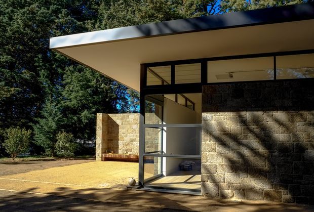Exterior view of a low-slung modern building set among mature trees. The composition is defined by a broad flat roof with a dark fascia, extensive black-framed glazing, and substantial warm-toned stone masonry walls. A glazed entrance corner and paved forecourt are visible, with strong dappled sunlight and shadows across the facade.