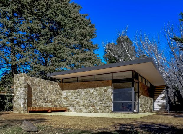 Single-storey modernist house set among mature trees, with extensive rough-cut stone masonry walls, dark timber or metal-framed clerestory glazing, a broad low-pitched projecting roof, and a glazed corner entrance. A timber bench runs along the stone wall, while a partially screened side area is visible at right. The composition emphasizes horizontal lines, simple geometric volumes, and an indoor-outdoor relationship.