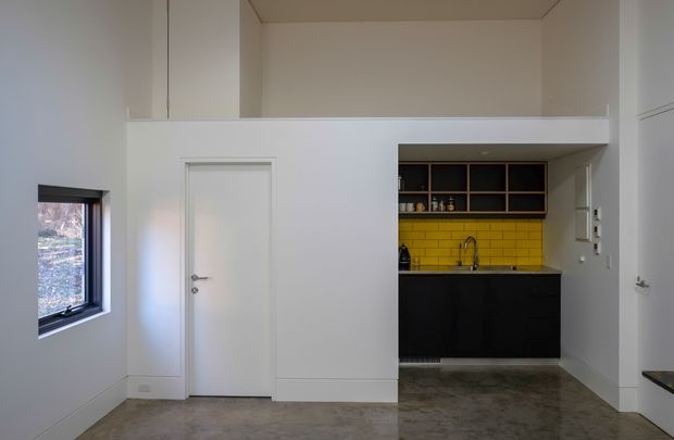 Compact open-plan interior with a minimalist white wall and concealed door enclosing a small kitchen area. The kitchen features matte black lower cabinetry, open dark timber shelving, a vivid yellow tiled splashback, and a stainless-steel sink. Polished concrete flooring, white walls, high ceilings, and dark-framed windows create a clean, modern industrial feel.