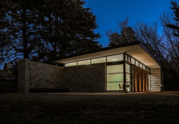 A low, single-storey modern pavilion photographed at dusk, set among mature trees. The building combines broad floor-to-ceiling glazing in dark frames with rough stone masonry walls, a thin projecting flat roof, and a sheltered glazed corner revealing a sparsely furnished interior. Warm interior and facade lighting contrasts with the deep blue evening sky and dark landscape.