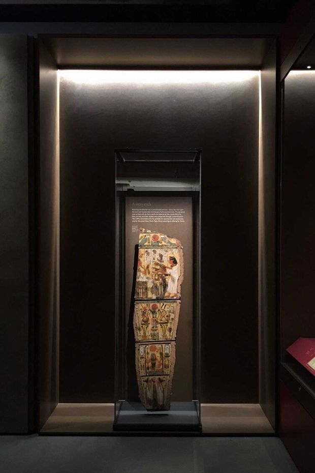 A dark museum or exhibition display featuring an illuminated glass-fronted case containing an ancient Egyptian decorated sarcophagus or coffin fragment. The display is set into a black architectural wall with concealed linear lighting, and a reflective glass panel and partial adjacent exhibit are visible at right.