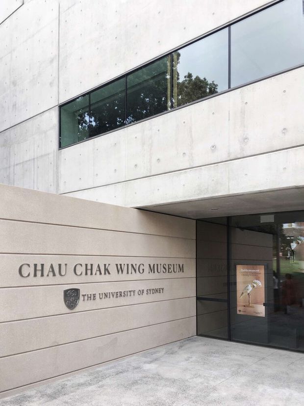 Exterior entrance of the Chau Chak Wing Museum at the University of Sydney. The modern institutional facade combines large smooth precast or poured concrete panels with visible circular form-tie marks, a long horizontal strip of dark framed glazing, and a recessed glass entry beneath a projecting concrete canopy. Museum and university signage is mounted on the pale stone-toned wall.