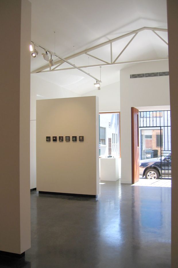 Bright open-plan gallery or studio interior with polished concrete flooring, white walls, exposed painted structural roof trusses, track lighting, and a large open doorway with security bars leading outside. A freestanding partition wall displays a row of small framed artworks or fixtures.