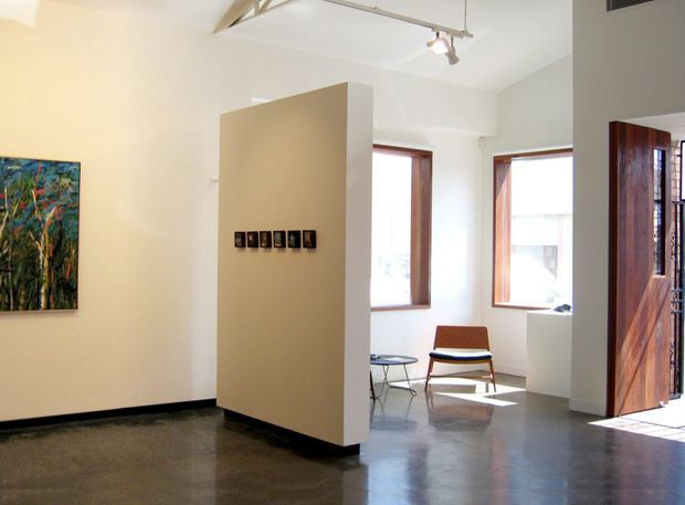 Bright contemporary interior with polished concrete flooring, white walls, exposed structural and service elements, timber-framed windows, and a large timber pivot door opening to the exterior. A freestanding partition wall displays small artworks, while a mid-century-style lounge chair and side table sit near the windows.