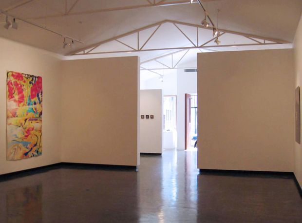A bright, open gallery-like interior with polished concrete flooring, white partition walls, exposed triangular roof trusses, and track lighting. Colorful abstract artwork is displayed on the left wall, while an open doorway and additional gallery spaces are visible beyond the partitions.