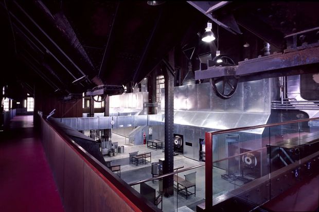 A dark, industrial-style commercial interior viewed from an elevated walkway or mezzanine. The space features exposed steel beams and columns, large metal ventilation ductwork, suspended industrial lighting, metal-clad walls, glass balustrades, and a lower-level open seating or dining area with tables and stools. Deep burgundy and black tones contrast with silver metal surfaces.