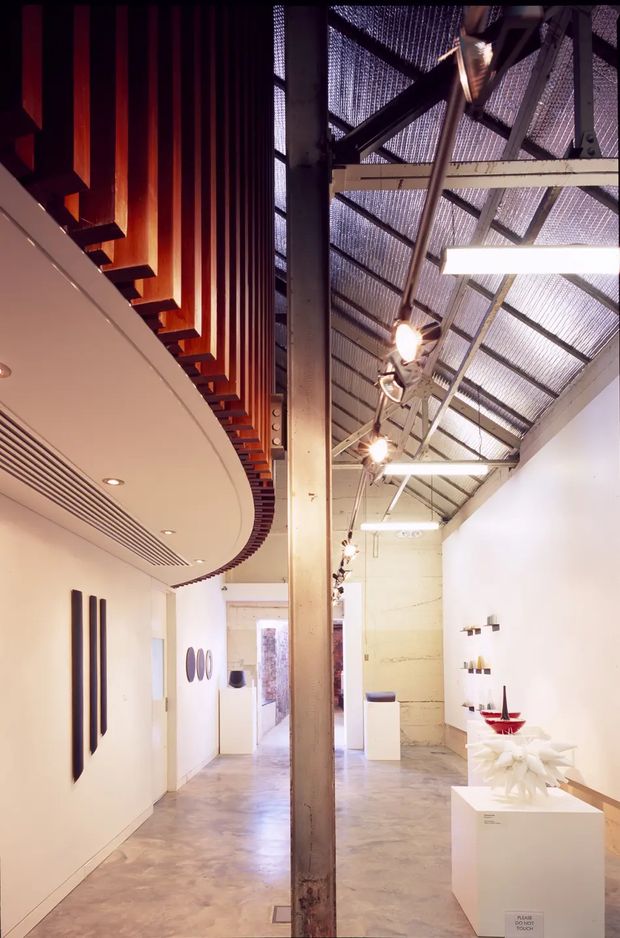 A tall, narrow contemporary gallery-like interior with polished concrete flooring, white walls, and exposed industrial roof structure. A central steel column divides the space, while dramatic timber slatted ceiling detailing curves along the left side. Track lights and linear fluorescent fixtures illuminate minimalist plinths and wall-mounted artwork or objects. The space has a bright, refined, adaptive-reuse atmosphere with strong vertical scale and exposed construction.