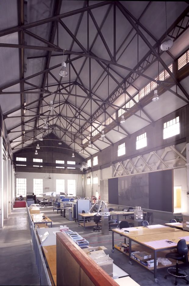 A large adaptive industrial-style interior resembling a converted warehouse or workshop, with an expansive pitched roof, exposed dark steel trusses, clerestory windows, high walls, suspended industrial lights, and rows of worktables and equipment. The space appears to function as a studio, workshop, or commercial creative workspace, with concrete flooring and a bright, utilitarian atmosphere.
