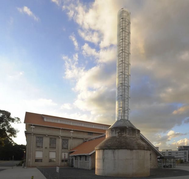 Exterior photograph of a substantial industrial or institutional building with a tall cylindrical concrete tower and enclosed metal framework rising above it. The main building has a pitched terracotta-colored roof, pale rendered walls, large multi-pane industrial windows, and exposed service or structural details. A paved forecourt and contemporary urban buildings are visible in the background beneath a partly cloudy sky.