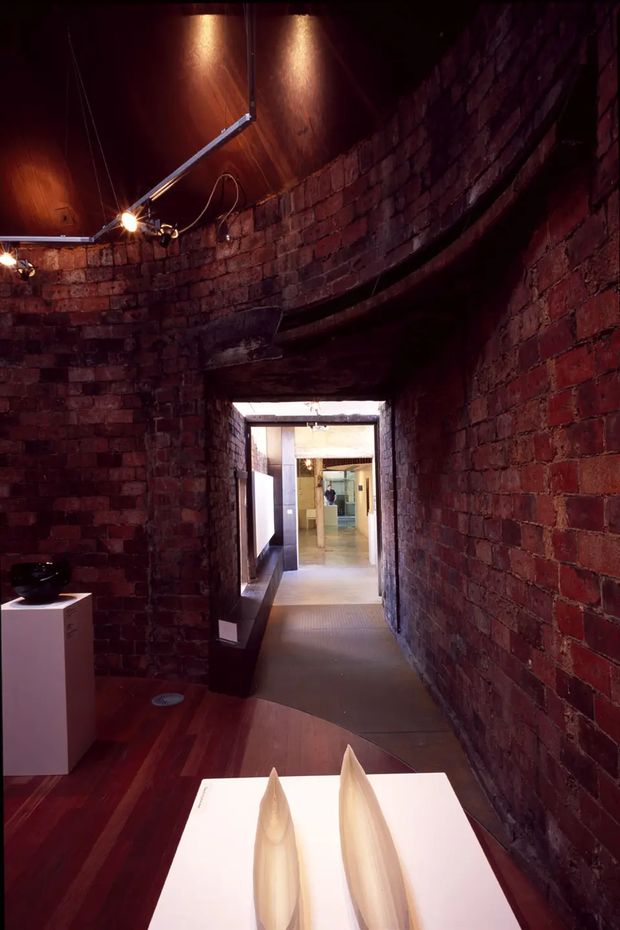 A dimly lit, narrow brick-lined interior passage or gallery with exposed red brick walls, a curved dark timber ceiling, and warm directional spotlights. A polished timber floor leads toward a brighter adjoining space through a rectangular opening. Minimal plinths and sculptural objects suggest an art gallery or exhibition setting.