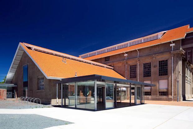 Exterior view of a substantial adaptive-reuse or heritage-style building with a newly added glazed entrance pavilion. The complex has large brick or masonry walls, tall industrial multi-pane windows, a prominent orange terracotta tiled pitched roof, and a modern dark-framed glass extension. The forecourt is broad and paved, with bicycle racks visible at the side.