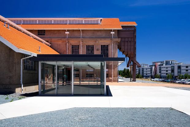 Exterior view of a heritage industrial or institutional building with terracotta tiled roofs, weathered masonry walls, tall multi-pane windows, and a contrasting modern glass entry pavilion. A paved forecourt and gravel area sit in front, with contemporary apartment or commercial buildings visible in the distance.