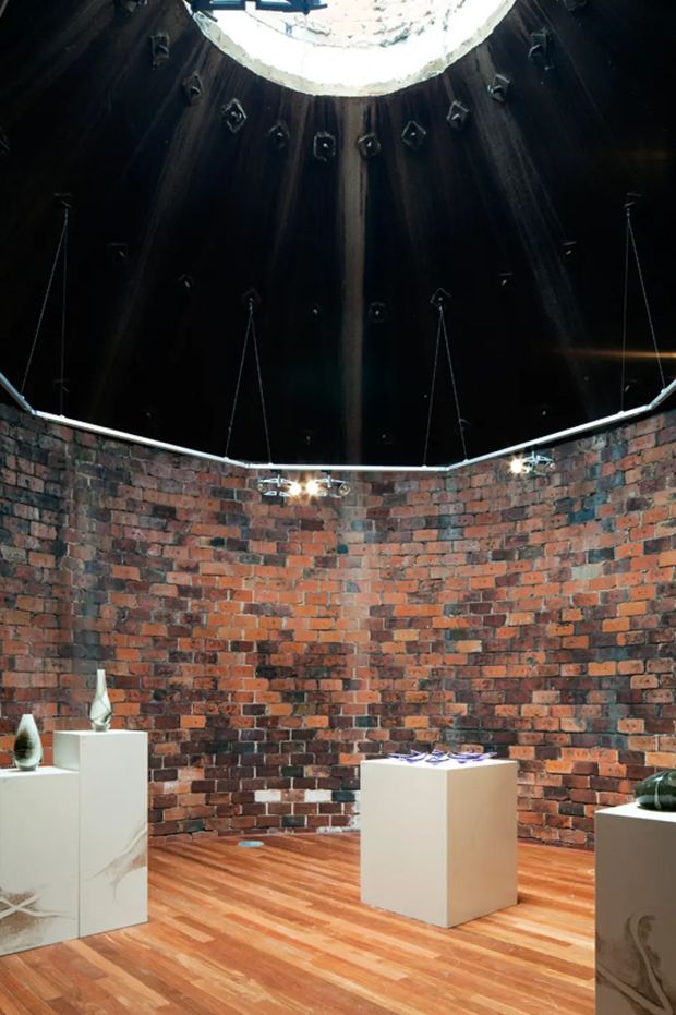 A circular gallery-like interior with exposed red and dark brick walls, a warm timber floor, white display plinths holding sculptural objects, and a dramatic dark domed ceiling. A large circular skylight or oculus at the apex casts beams of daylight into the space, while suspended track or pendant lighting follows the curved perimeter. The atmosphere is dramatic and industrial, with strong contrast between the masonry, dark ceiling, and bright display furniture.