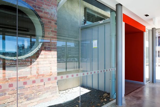 A contemporary commercial or institutional building entrance with a glazed façade, exposed red brick wall, large circular window, corrugated metal panel, and cylindrical metal columns. A bold red recessed doorway or corridor is visible to the right, with polished concrete flooring and strong natural light.