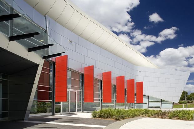 Exterior view of a contemporary curved building with a white panel-clad facade, sweeping roofline, extensive glazing, projecting red vertical sun-shading panels, and a paved landscaped forecourt. The composition emphasizes the building envelope and dramatic sculptural form.