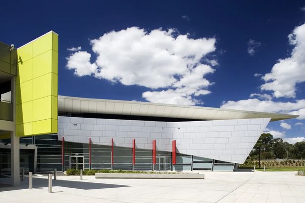 Exterior view of a contemporary commercial building with a sculptural white tiled facade, sweeping curved roof canopy, extensive glazing, vivid lime-green vertical feature wall, and red structural accents. A broad paved forecourt and landscaped grounds surround the building under a bright blue sky.