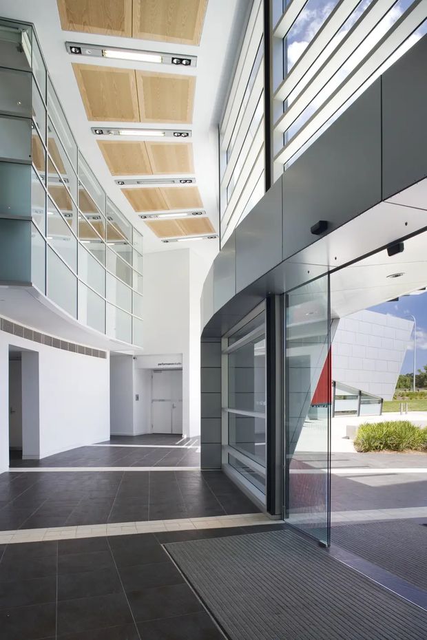 A bright contemporary commercial entrance or lobby with a tall glazed facade, dark charcoal tiled flooring, white walls, and large dark-framed glass doors. The high ceiling features repeated light timber acoustic panels with integrated linear lighting, while a curved glazed mezzanine or upper-level wall lines the left side. Exterior landscaping and a sculptural white building element are visible through the glazing.