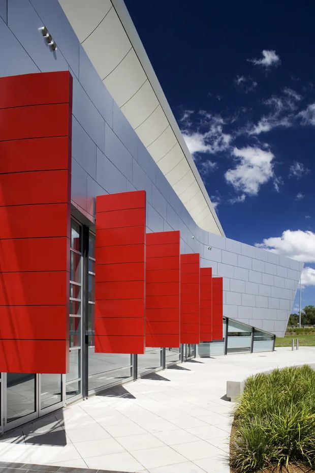 Exterior view of a contemporary commercial building with a sweeping curved roof edge, pale grey metal panel cladding, extensive glazed doors and windows, and a row of bold red vertical facade fins. A paved forecourt and landscaped planting sit alongside the building under a bright blue sky.