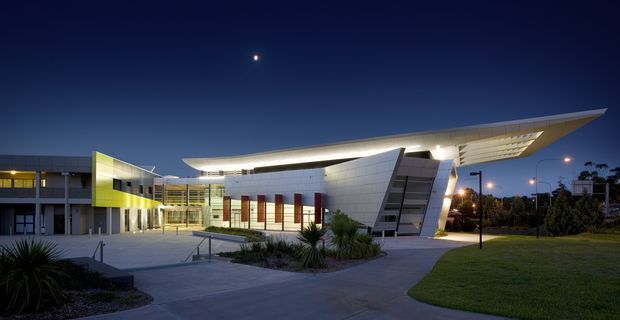 Twilight exterior view of a contemporary institutional or commercial building. The complex features angular white-clad volumes, a dramatic sweeping roof canopy, extensive glazing, illuminated interiors, and a broad paved forecourt with landscaped planting and lawn.
