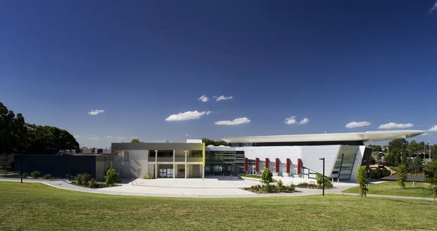 Wide exterior view of a contemporary institutional or commercial building set within landscaped grounds. The complex combines low, rectilinear white and grey volumes with extensive glazing, a prominent angular projecting roof or canopy, and a series of vivid red vertical facade elements. A paved forecourt, lawn, young trees, and clear blue sky surround the building.