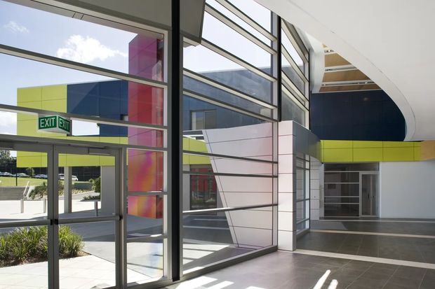 Bright contemporary commercial interior lobby or entry space with extensive floor-to-ceiling glazing, dark metal window frames, polished grey tiled flooring, and a striking curved white ceiling. Bold lime green, navy blue, magenta, and white architectural panels are visible through the glazing and along the upper walls. An exit door opens toward landscaped exterior grounds.