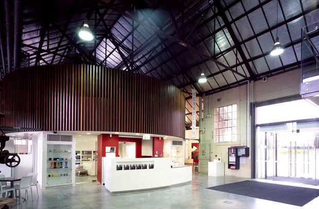 A spacious industrial-style commercial reception or entry area with a large white curved reception desk, exposed dark steel roof trusses, corrugated metal ceiling, high clerestory windows, polished concrete flooring, and a dramatic curved wall clad in vertical timber slats. Bright pendant lights and expansive glazed entry doors contribute to the open, functional space.