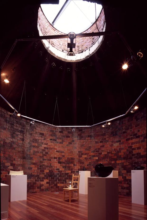 A dark circular brick-walled exhibition or gallery interior with timber flooring, white display pedestals, sculptural artworks, and focused track lighting. A large circular skylight or roof opening with exposed brickwork and structural framing brings light into the centre of the space.