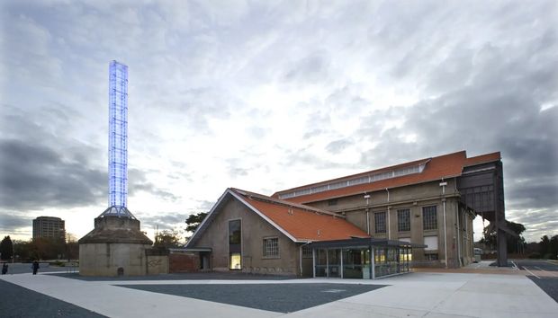 Exterior view of a substantial civic or industrial-style building complex under a dramatic cloudy sky. The foreground shows a broad paved plaza, with a low gabled masonry building featuring a red tiled pitched roof and glazed entrance attached to a taller monumental structure with large multi-pane windows, exposed stone or concrete detailing, and a dark projecting upper volume. A tall narrow illuminated or glazed tower rises beside the complex.