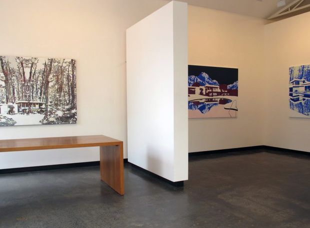 A minimalist gallery-like interior with pale beige walls, a polished dark grey concrete floor, black skirting, and a freestanding white partition wall. Several framed artworks are displayed on the walls, while a simple timber console table occupies the foreground. Exposed ceiling services and a linear overhead element contribute to the commercial exhibition feel.
