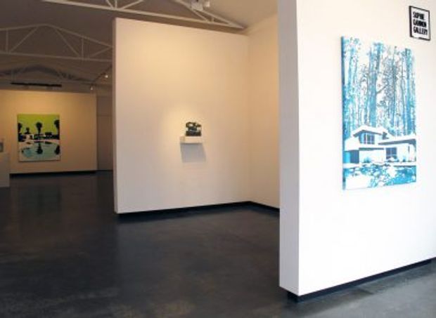 A minimalist gallery interior with white partition walls, dark polished concrete flooring, exposed white-painted ceiling trusses, and sparsely displayed contemporary artworks. A small sculpture sits on a wall-mounted shelf, while framed blue-toned artwork is mounted on the foreground wall. The space is open, quiet, and industrial in character.