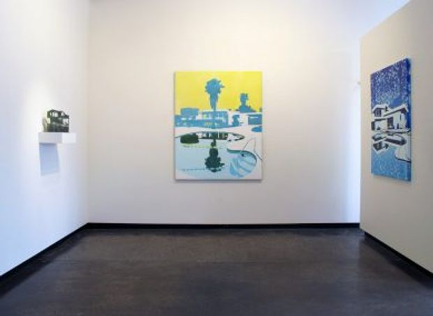 Minimal gallery-like interior with white walls, dark polished concrete flooring, black skirting, and colorful blue-and-yellow artworks mounted on the walls. A small white wall shelf with a decorative object is visible on the left. The space is open, sparse, and likely a commercial exhibition or gallery area.
